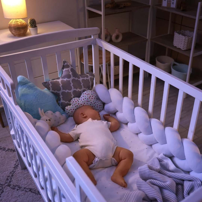 A baby doll lying on a white crib mattress surrounded by white crib bedding, including a braided bumper pad, plush pillows, and a striped blanket, in a softly lit nursery.
