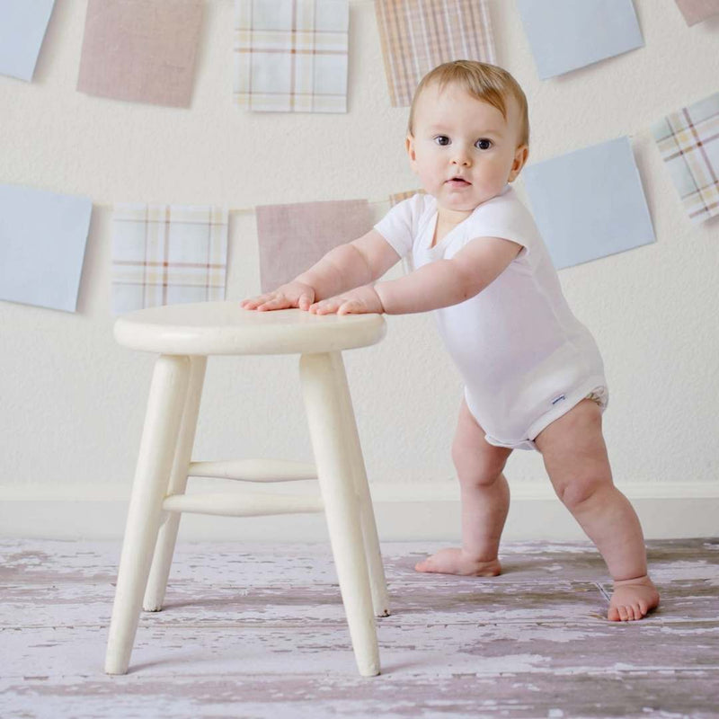 A baby wearing a white bodysuit stands on a distressed wood floor, balancing on one foot and pushing a small cream-colored stool. The background shows a cream wall decorated with fabric squares and plaid banners.