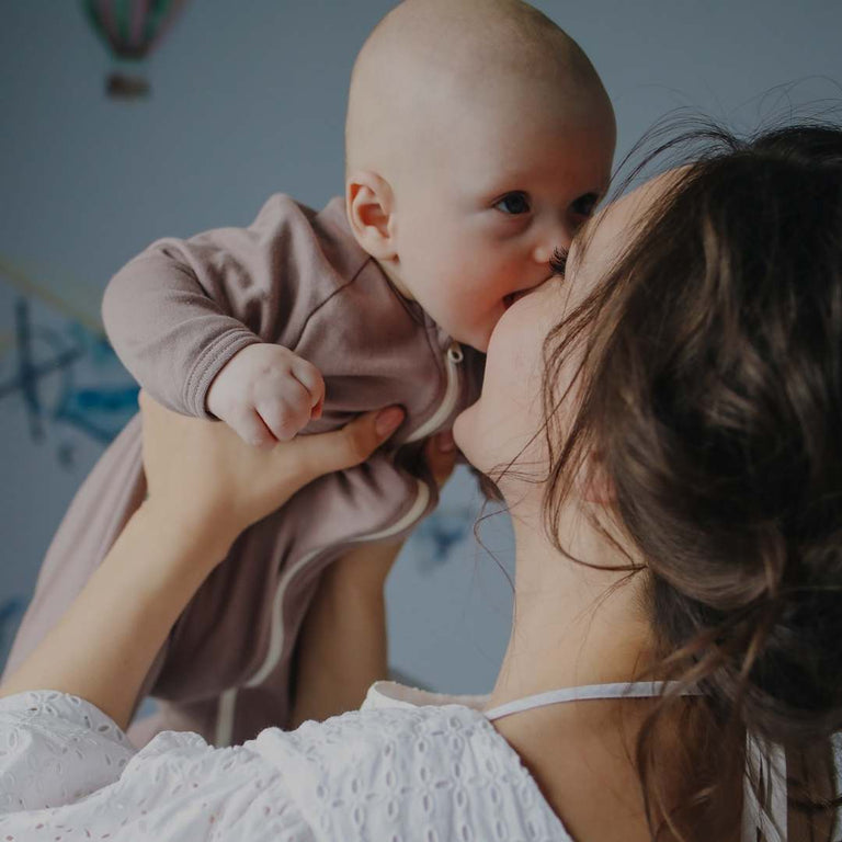 A woman in a white lace-trimmed top holds and kisses a baby wearing a dusty mauve-pink long-sleeve bodysuit. The photo is taken in a softly lit room with a blue-gray wall and subtle wall decals in the background.