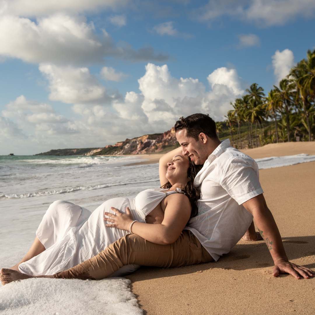 A couple in white and tan clothing sitting together on a sandy beach with the ocean, clouds, and palm trees in the background.