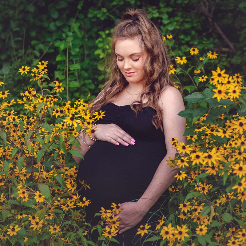 A pregnant woman in a black strapless dress stands in a field of yellow flowers, cradling her baby bump. She has long wavy light brown hair and is wearing a delicate gold necklace and bracelet.