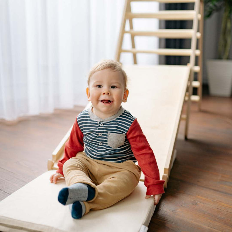 A baby sitting on a wooden toddler slide in a bright room with wooden floors and white curtains.