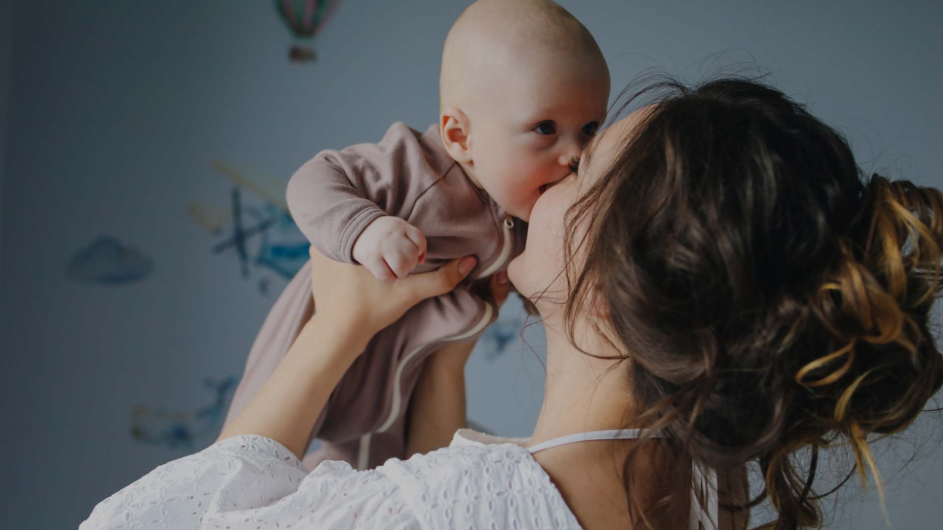 A woman in a white lace-trimmed top holds and kisses a baby wearing a dusty mauve-pink long-sleeve bodysuit. The photo is taken in a softly lit room with a blue-gray wall and subtle wall decals in the background.